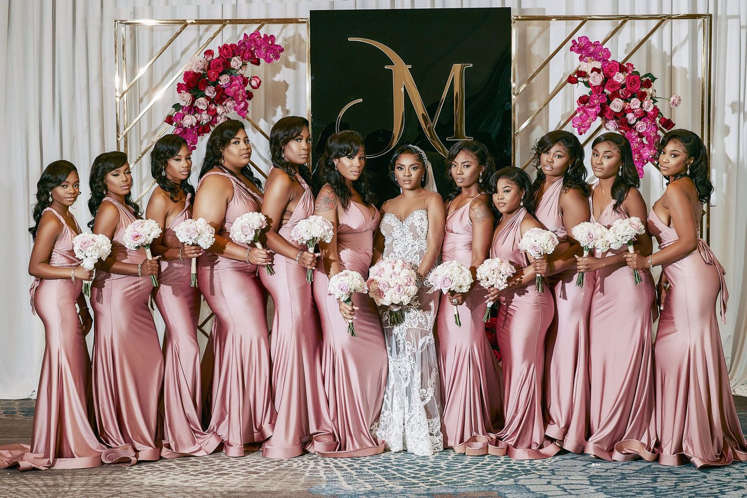 Bride with bridesmaids in pink satin mermaid dresses holding white bouquets at elegant wedding venue.