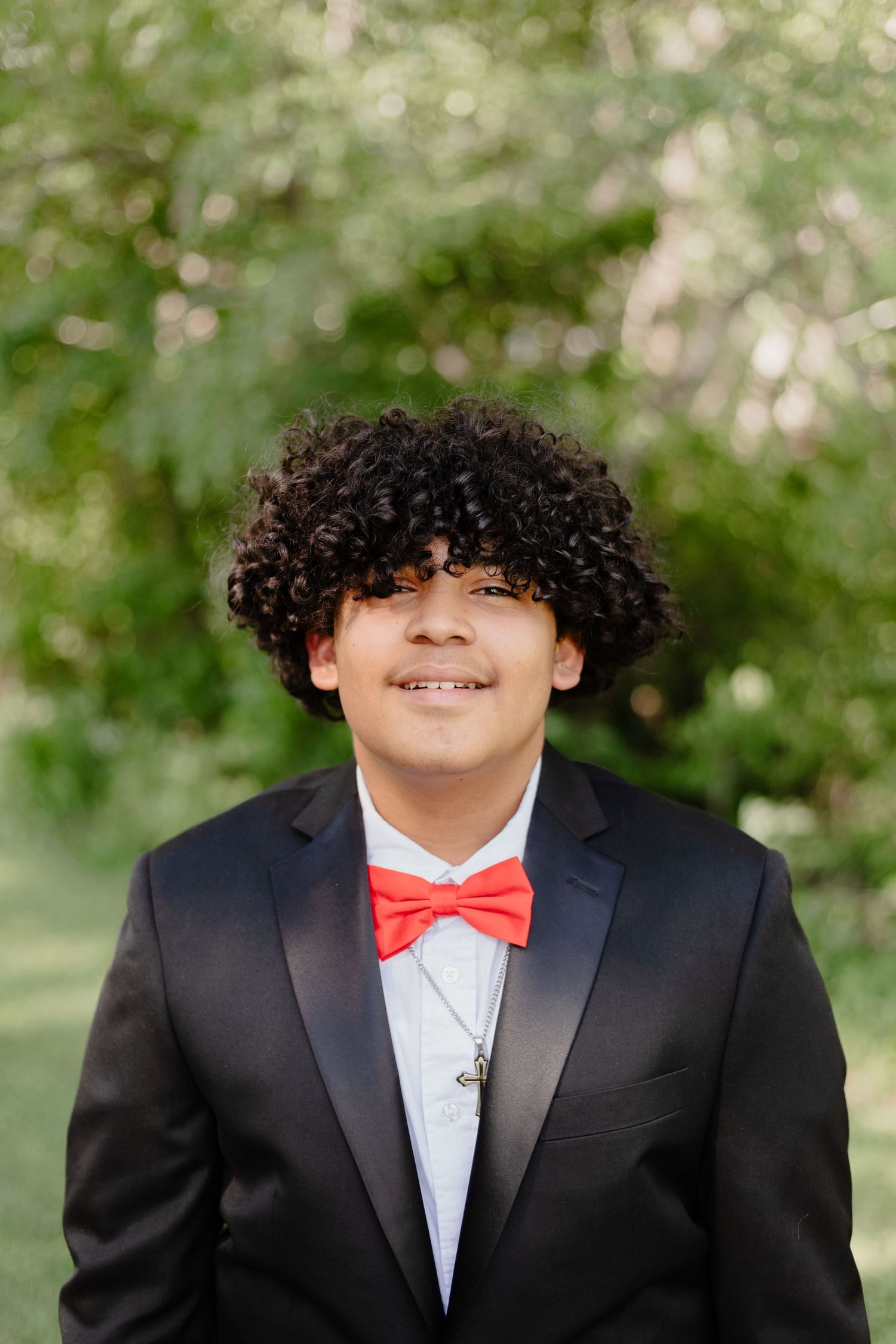 A formal portrait of someone wearing a black tuxedo and red bow tie against a nature background.