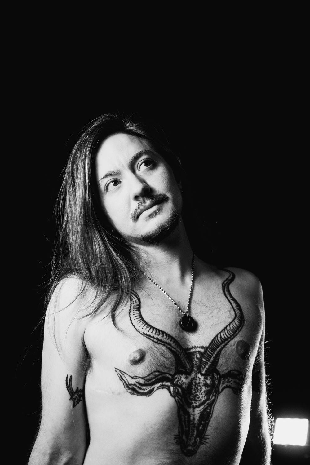 Black and white portrait of shirtless man with long hair and goat skull chest tattoo.