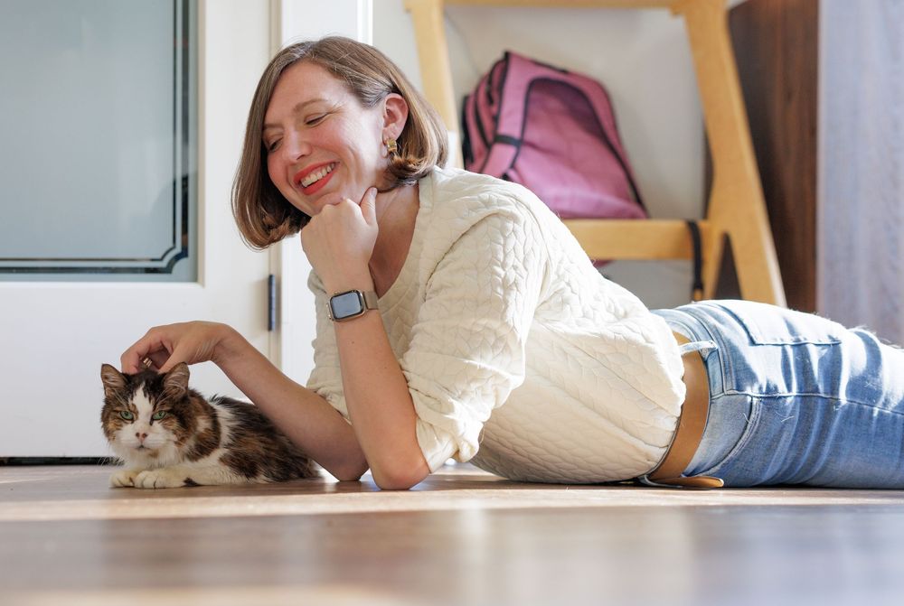 Young woman lying on the floor smiling while petting a small cat in a cozy indoor setting.