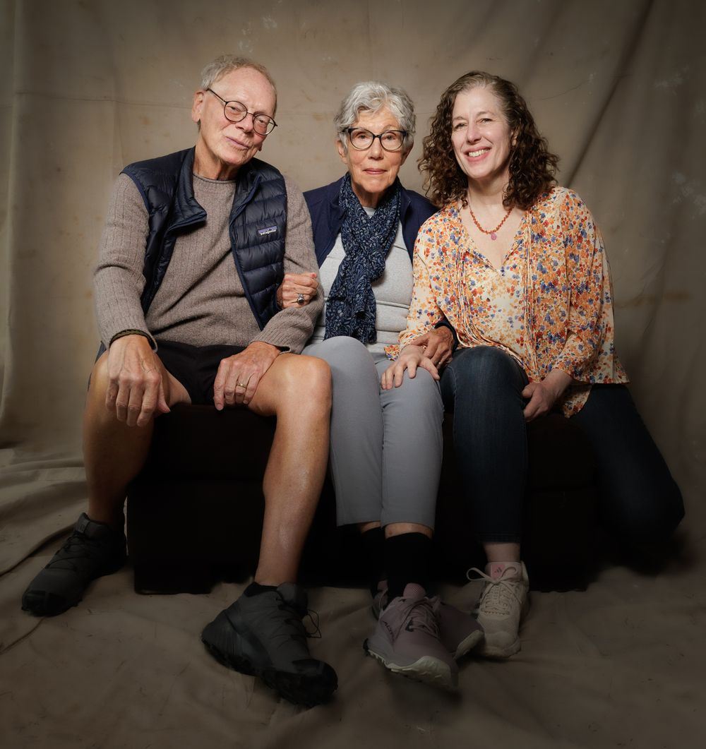 Three adults sitting together smiling in casual attire against neutral studio background.