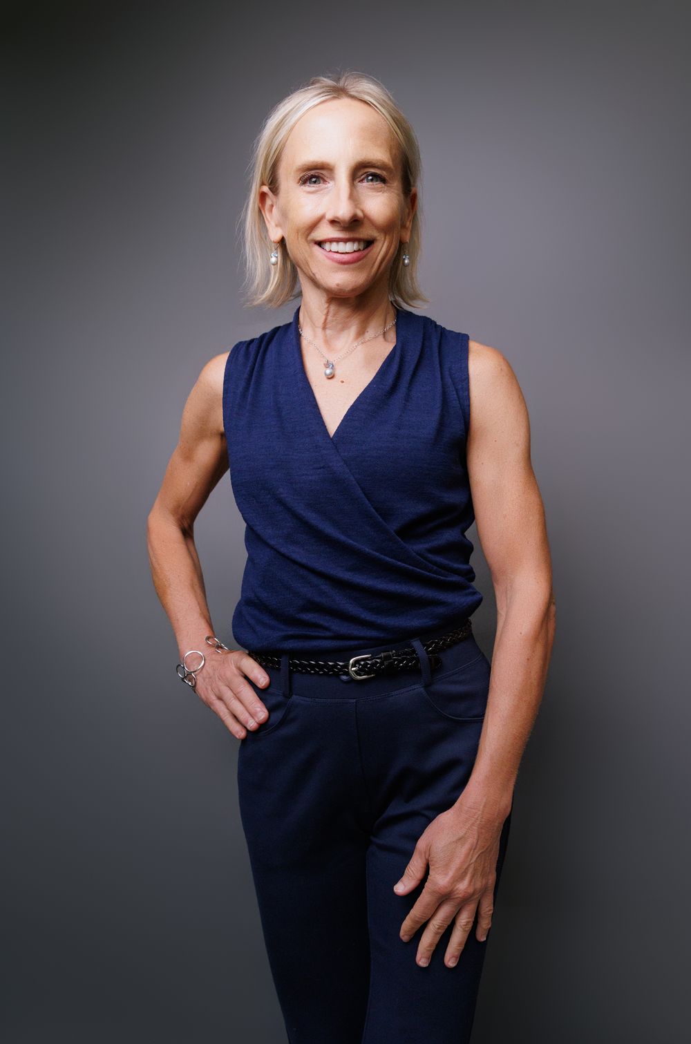 Confident woman in sleeveless blue top and navy pants posing against gray studio background.