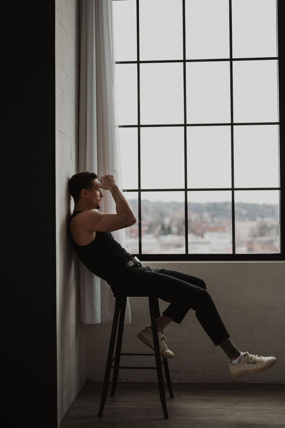 Artistic silhouette photo of person sitting on stool by large window in moody lighting.