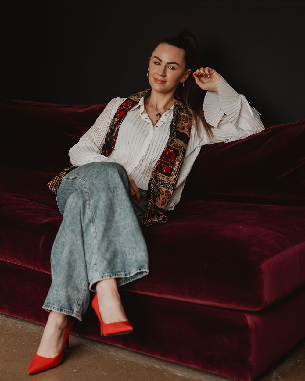 A person in a white shirt with a vintage vest lounges on a red velvet couch wearing red heels and light denim jeans.