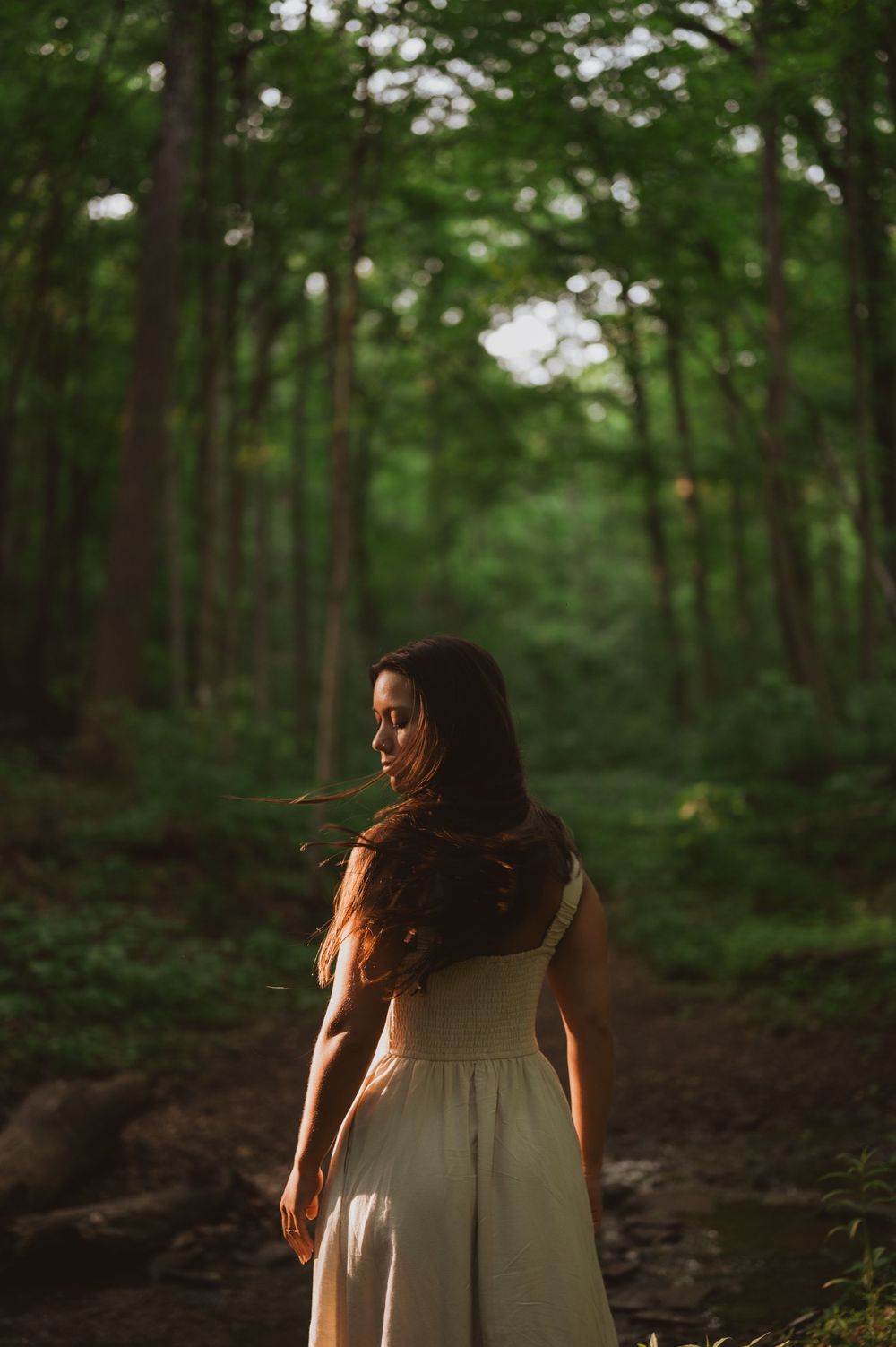 A white sundress-clad figure stands in a sunlit forest path surrounded by lush green trees.