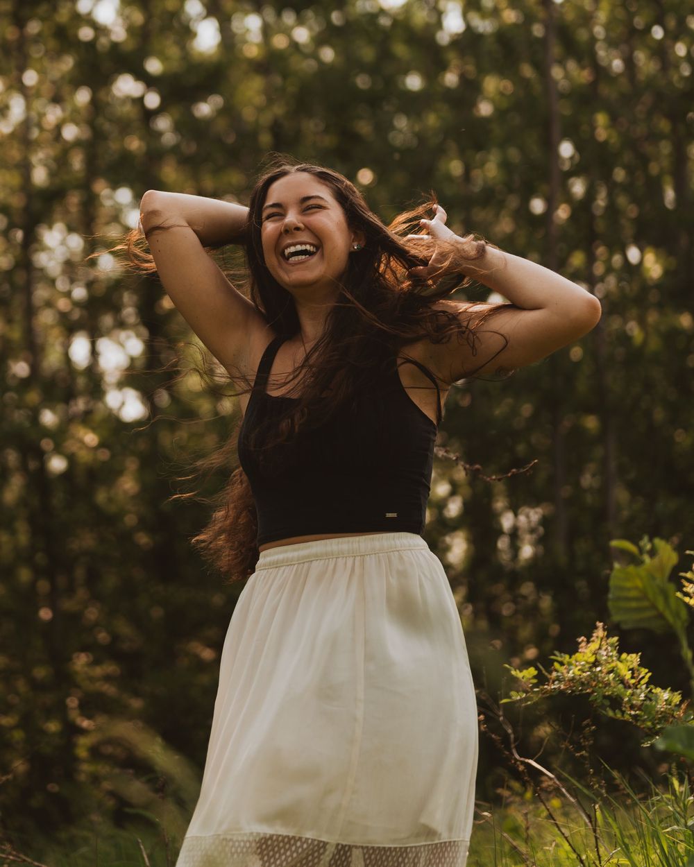 A joyful person in a black top and white skirt dances freely in a sunlit wooded area.