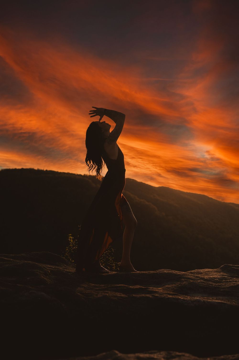 A silhouetted figure stretches against a dramatic orange and red sunset sky over mountain ridges.