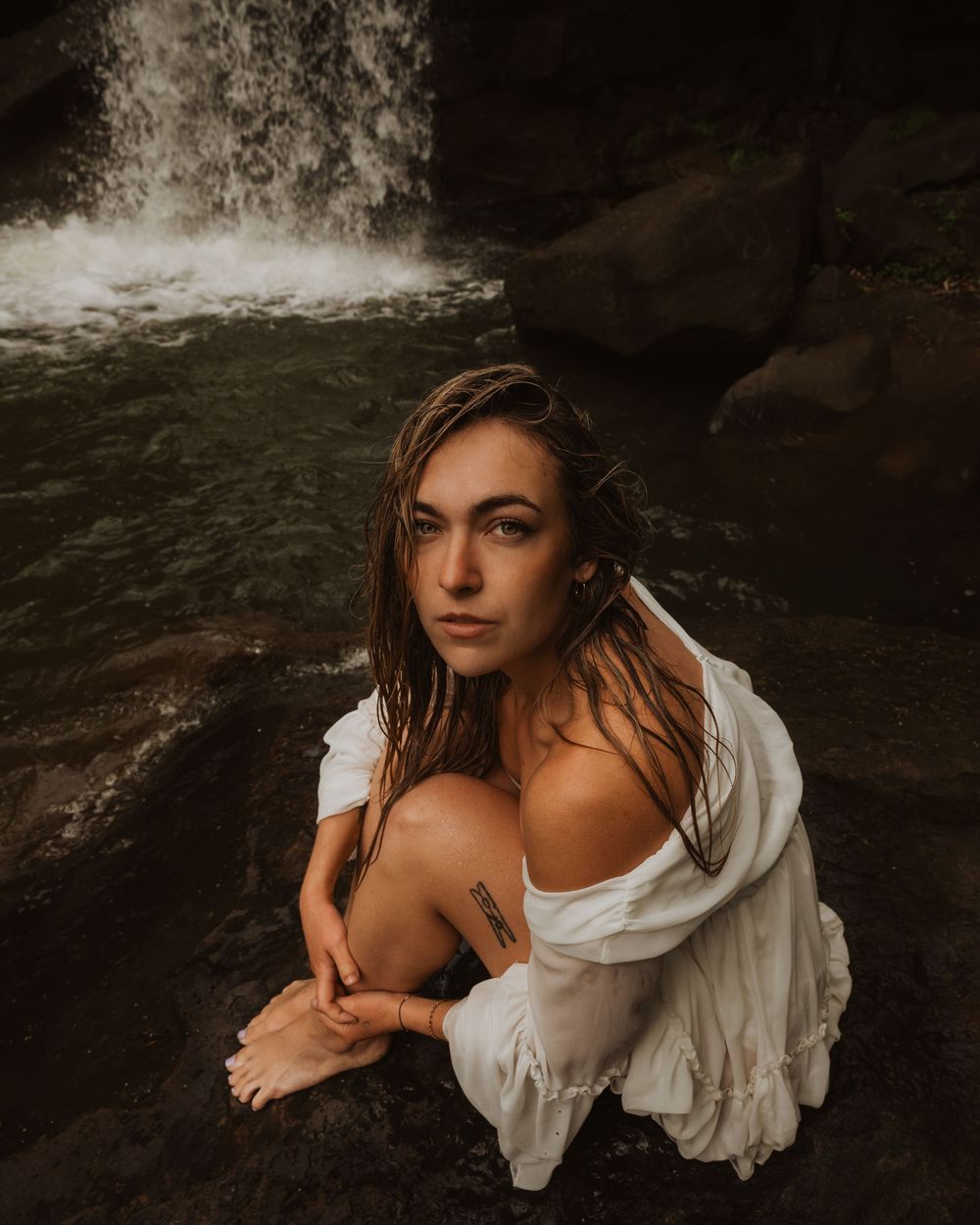 Dark and moody fine art portrait series of a woman in a white dress posing in flowing water against natural rock formations.