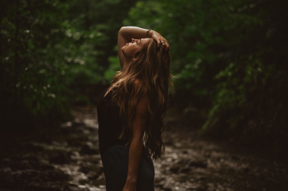A person with long brown hair walks along a wooded forest path surrounded by lush green vegetation.