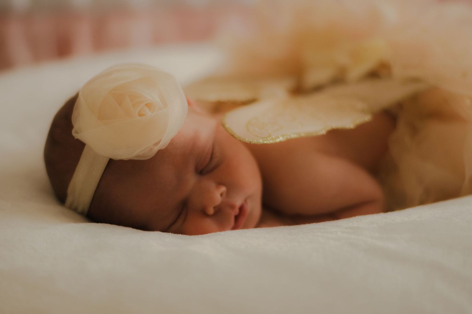 Sleeping newborn baby wearing a white headband rests peacefully on soft beige bedding in warm lighting.