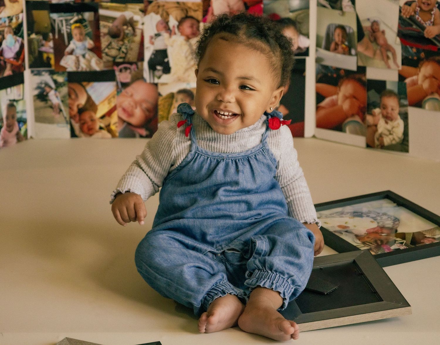 Adorable smiling baby in denim overalls sits surrounded by framed family photos on white background.