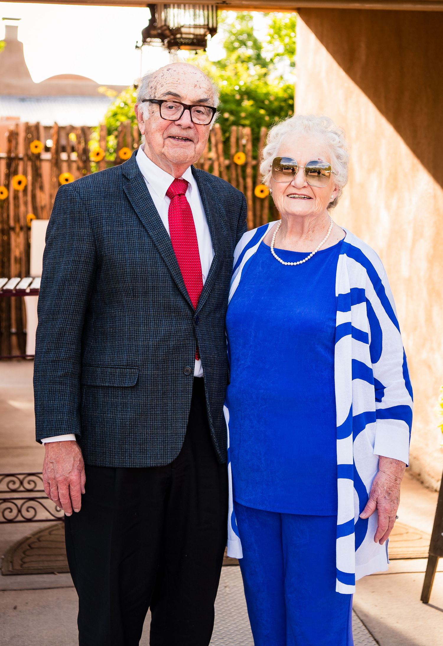 Elderly couple in formal attire, with one in a navy suit and red tie and the other in a bright blue outfit.