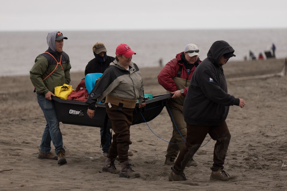 Group of people carrying equipment across a beach in cold weather gear.