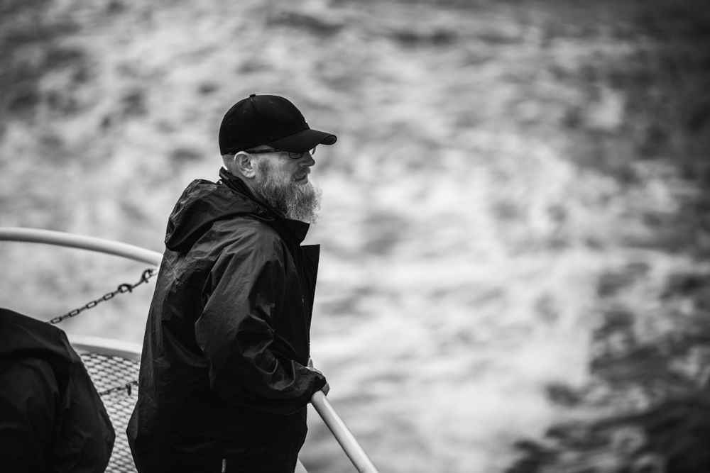 A black and white photo of someone in a dark jacket and cap standing on a boat.