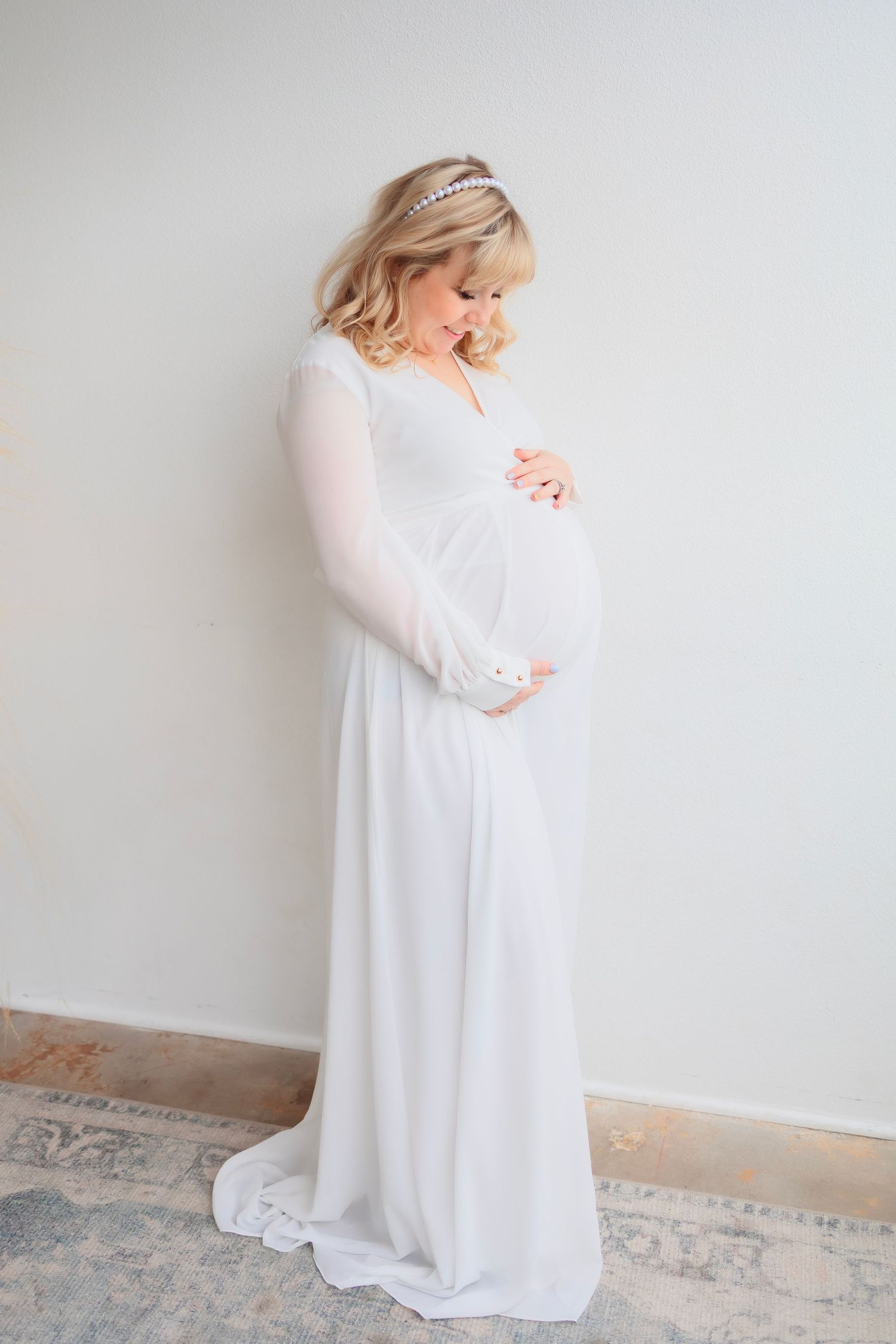 Pregnant woman in flowing white maternity dress stands in profile against white background displaying baby bump in studio setting.