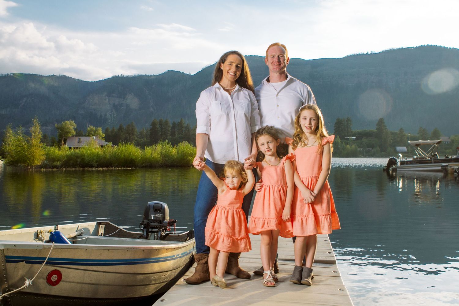 A family stands together on a wooden dock by a lake with mountains in the background and a boat nearby.