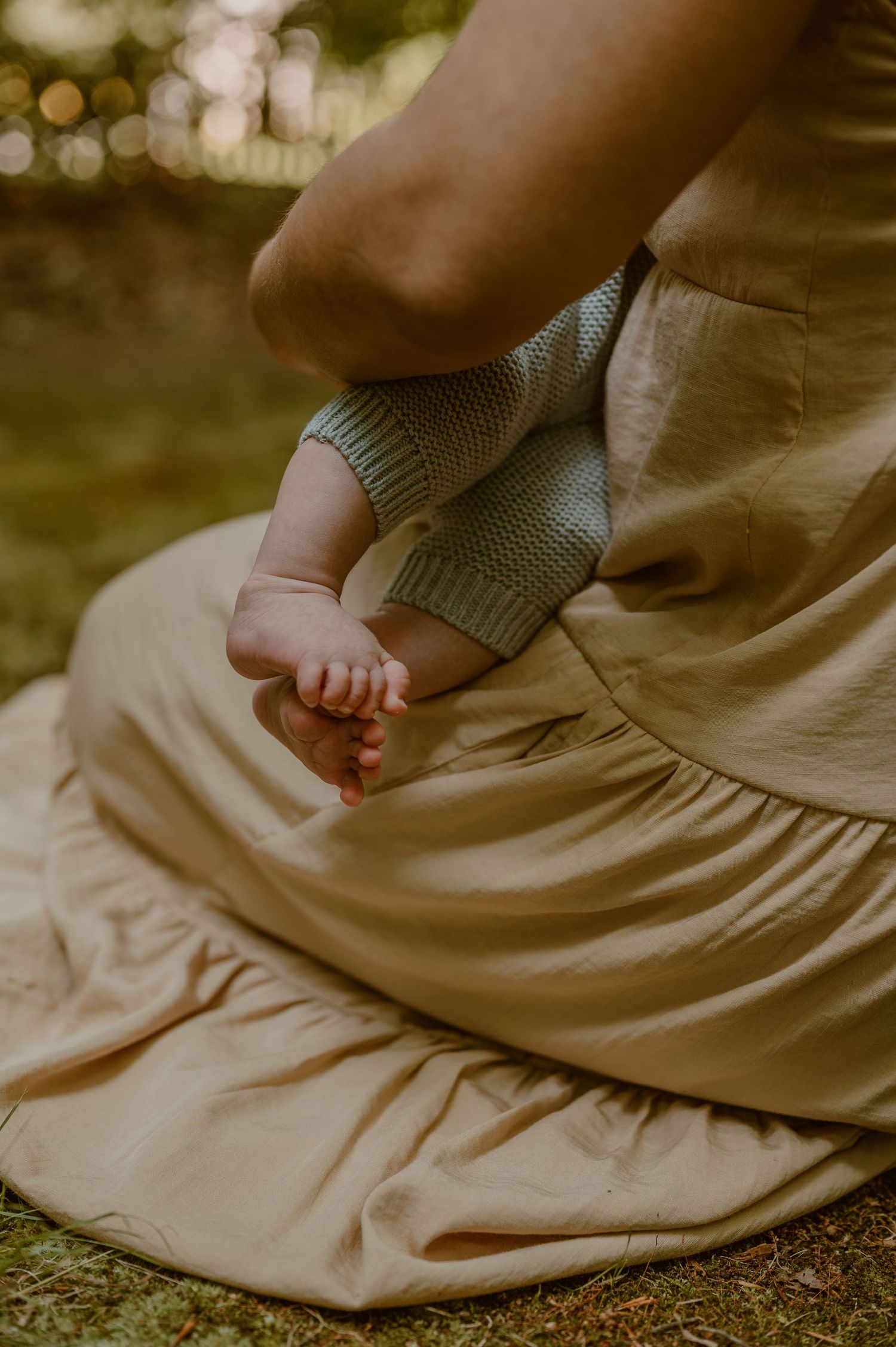 A tiny baby foot rests against a sage green knit sweater while sitting on a beige flowing dress outdoors.