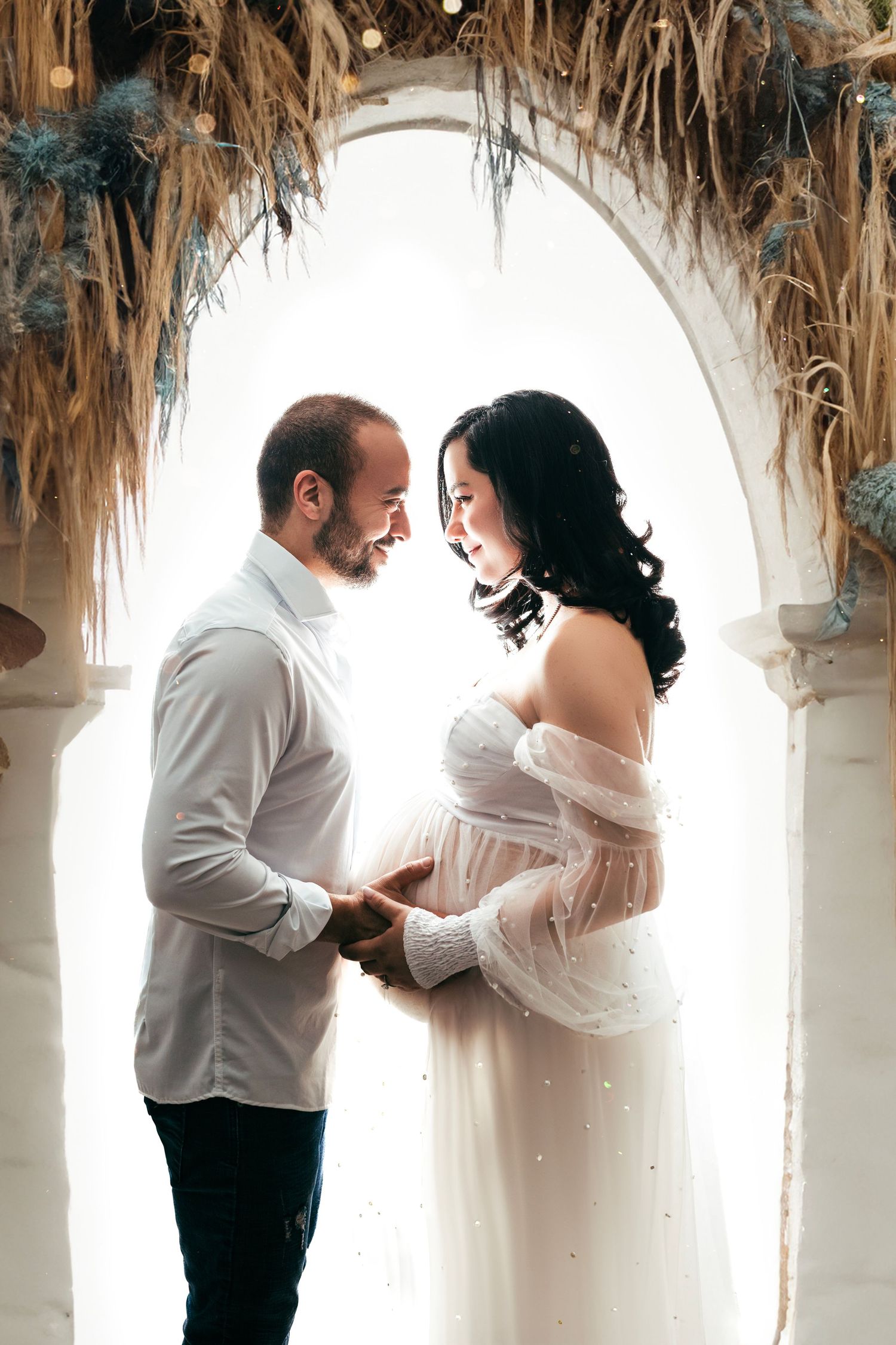 Romantic maternity photo of couple in white under rustic arch with dried flowers.