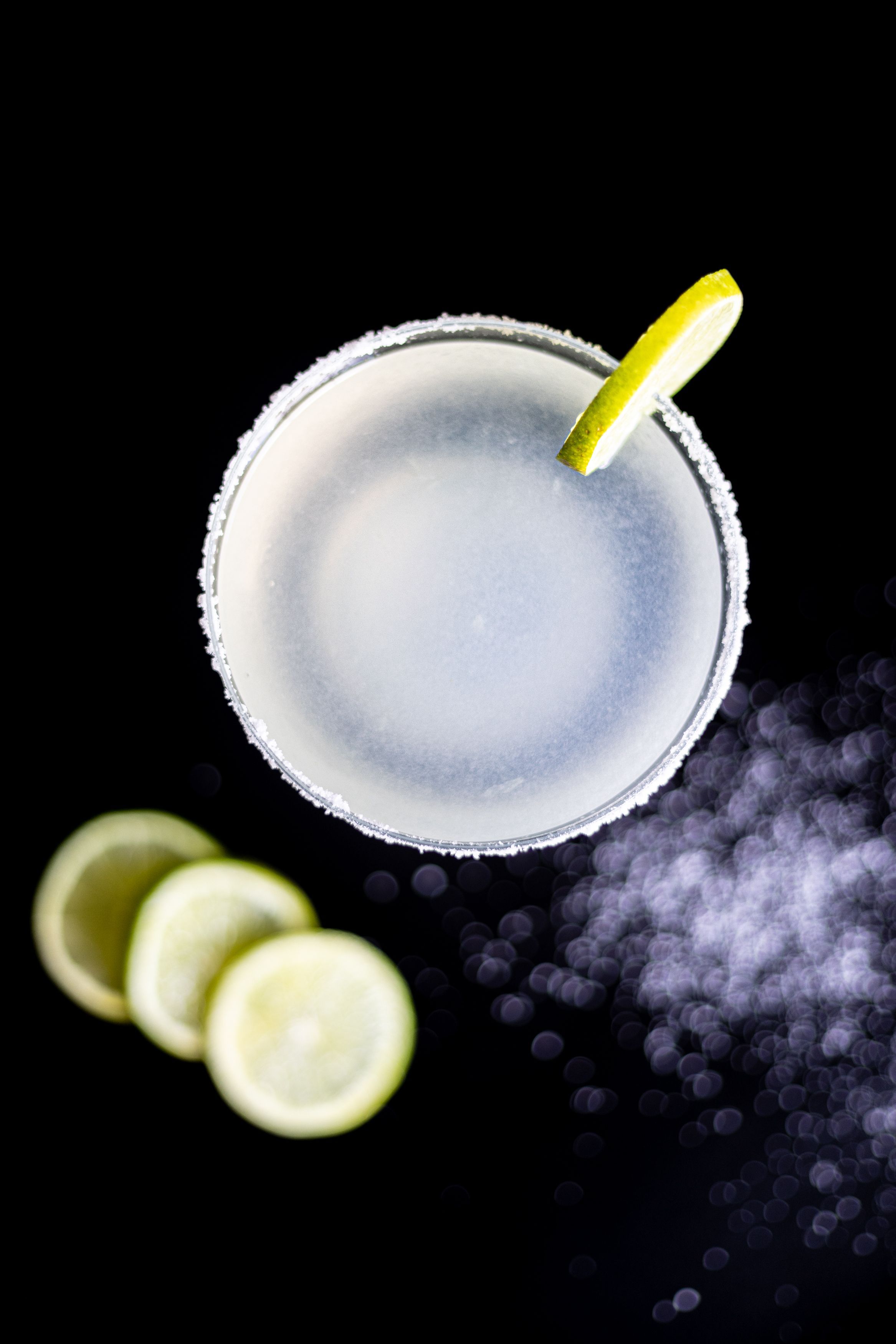 White cocktail with salt rim and lime garnish viewed from above on dark background.