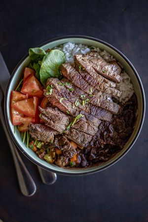 Rice bowl topped with sliced grilled steak, tomatoes, and fresh garnishes.