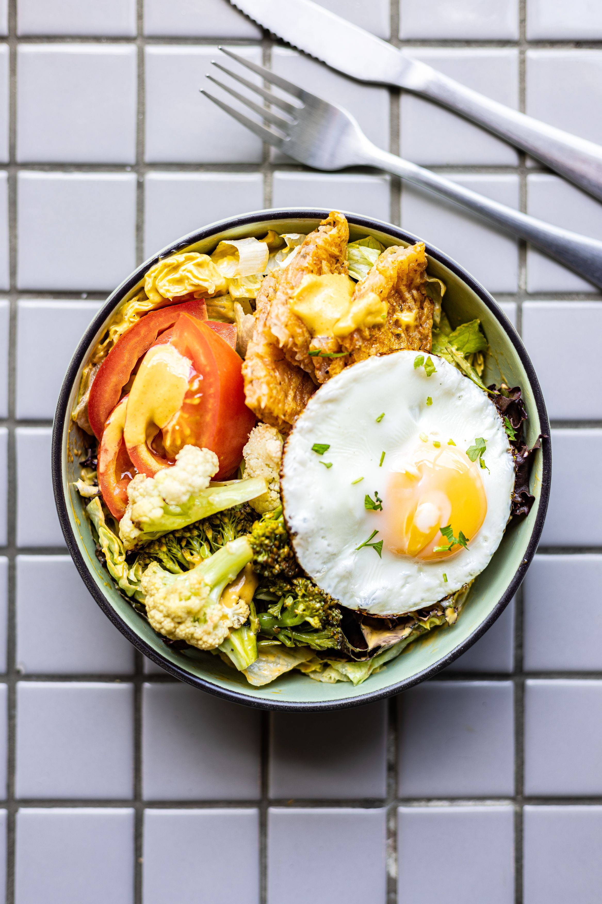 Fresh breakfast bowl with sunny side up egg, tomatoes, and mixed vegetables on tiled surface.