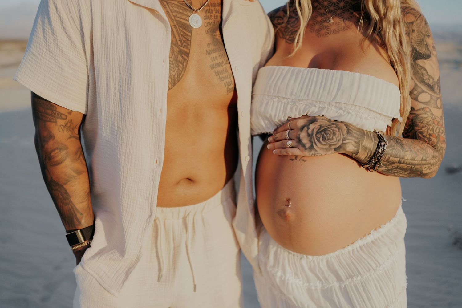 Close-up of tattooed couple in white clothing during desert maternity shoot.