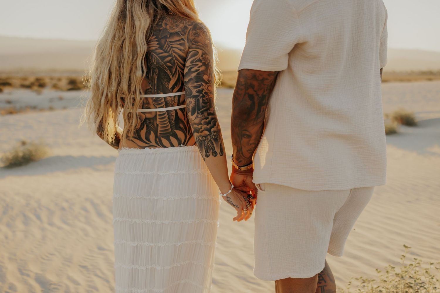 Couple holding hands in white outfits on a desert landscape during golden hour.