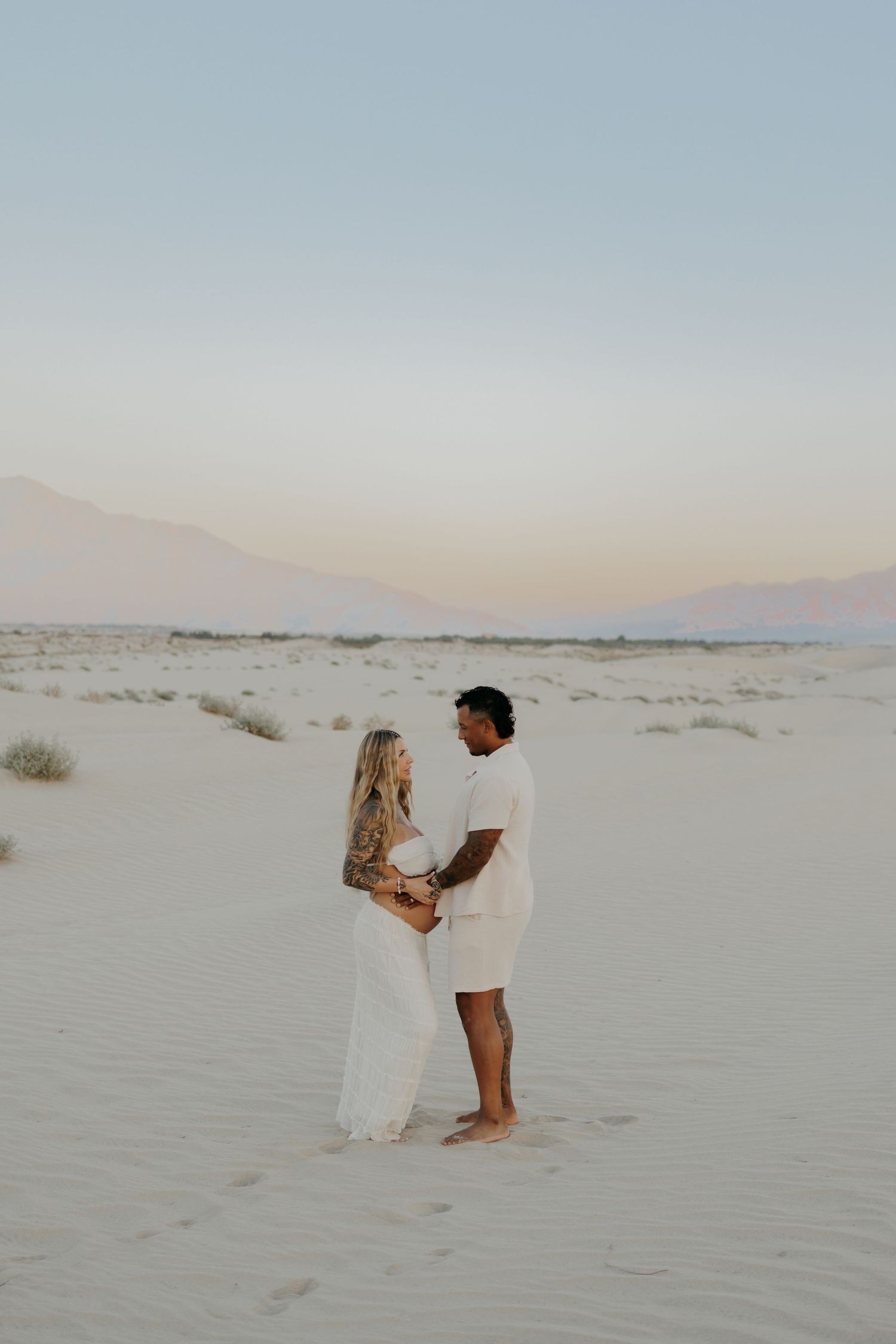 Two people share an intimate moment standing on white sand dunes at dusk.