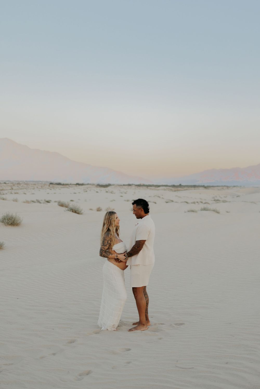 Two people share an intimate moment standing on white sand dunes at dusk.