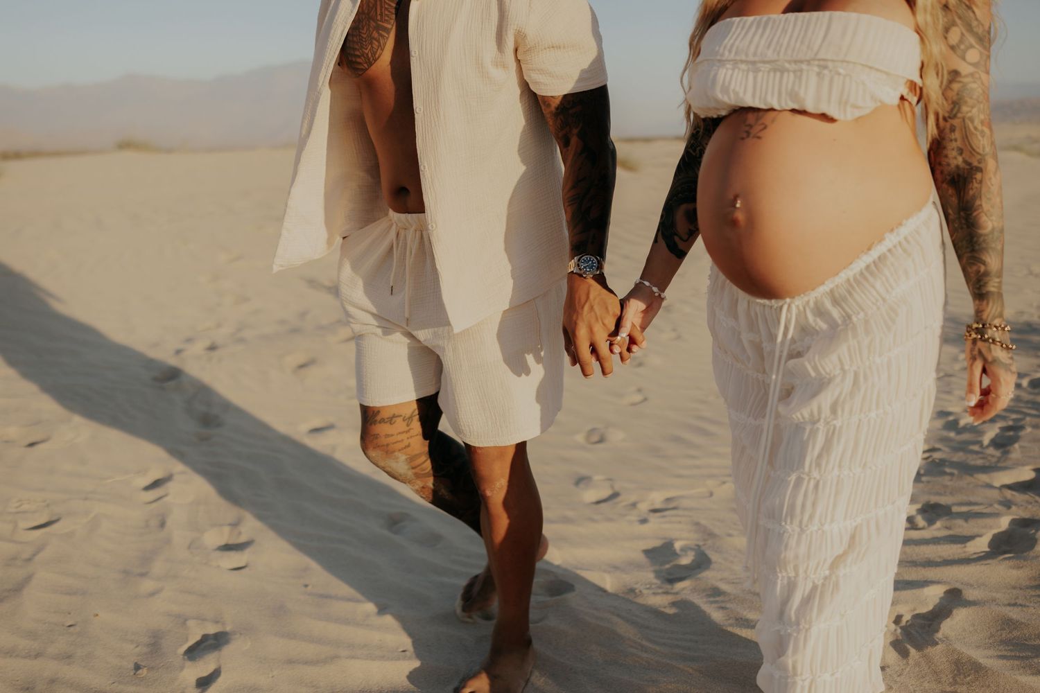 Close up of couple holding hands while walking in sand wearing flowing white clothing.
