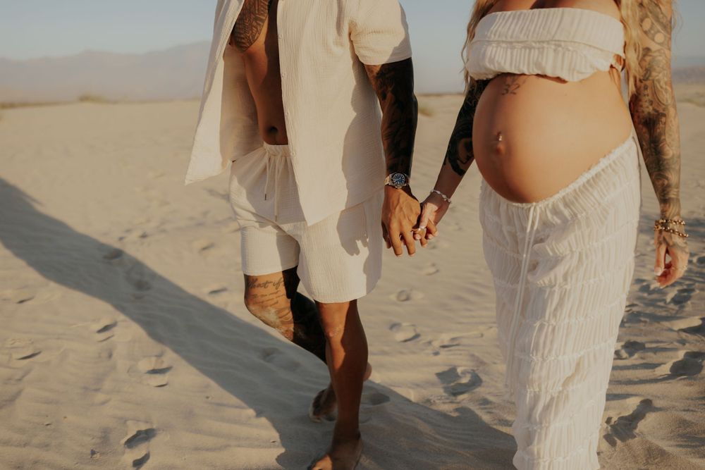 Close up of couple holding hands while walking in sand wearing flowing white clothing.
