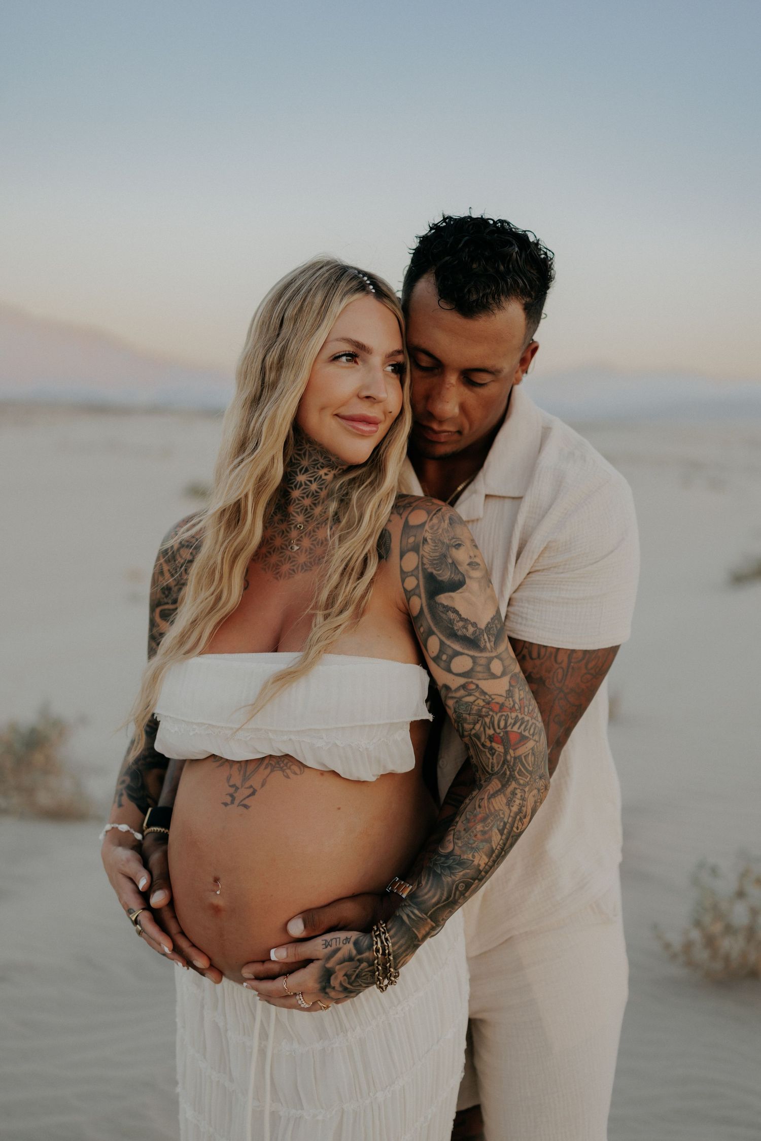 Maternity photo of couple in white embracing in desert setting during golden hour.
