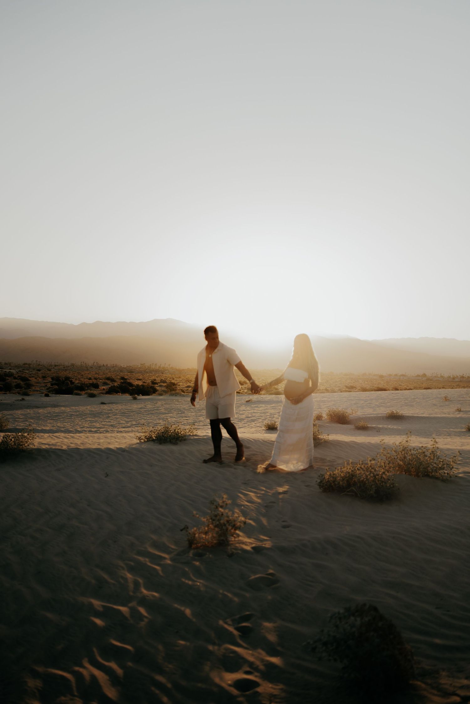 Couple in white outfits holding hands while walking on sand dunes during golden hour sunset.