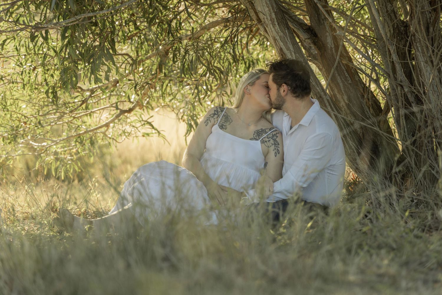 A couple shares an intimate moment under tree branches during a sunset photo session.