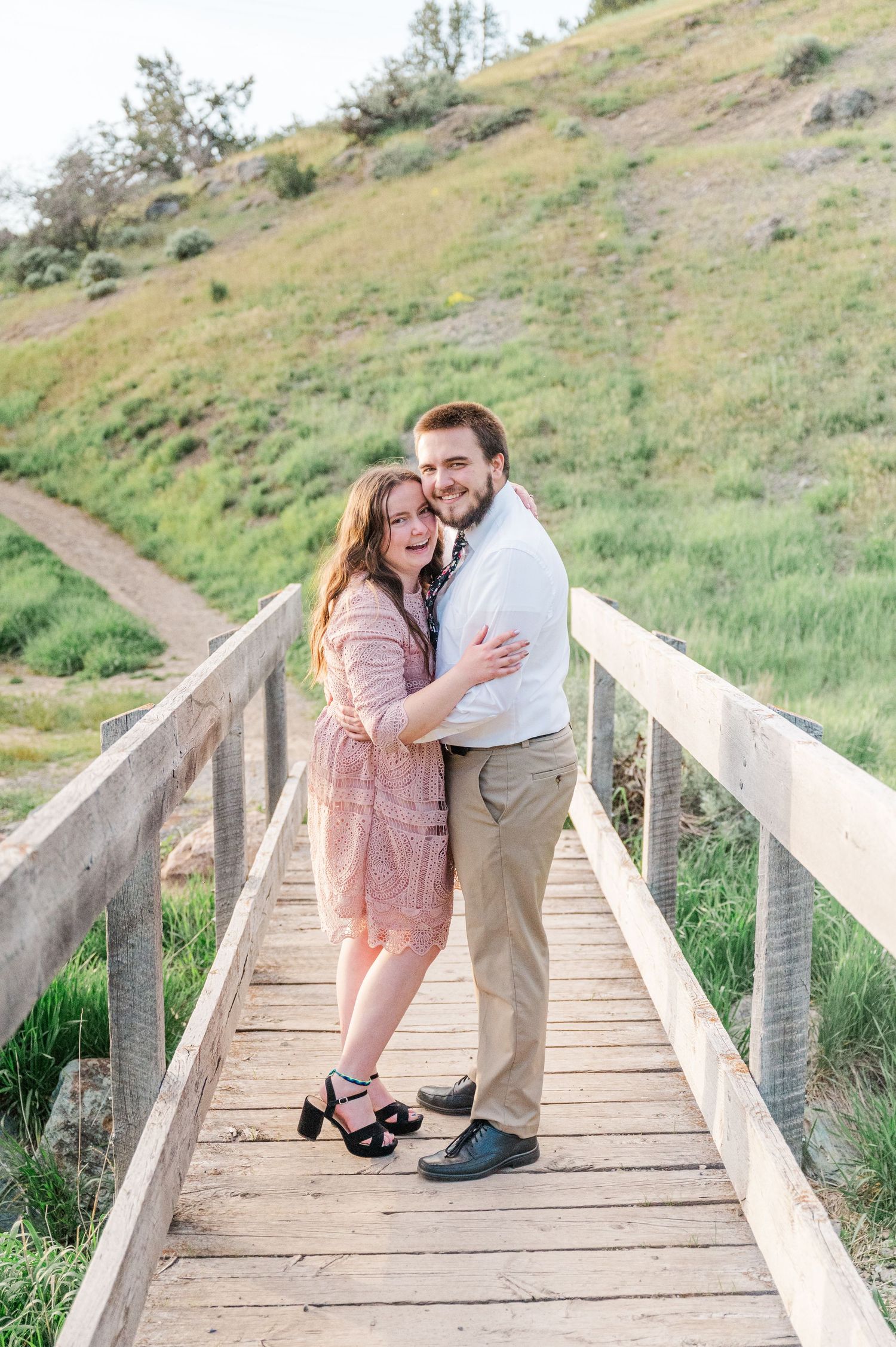A couple embraces on a wooden bridge with green hills and nature in the background.
