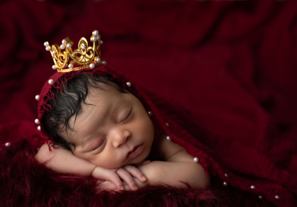 Sleeping newborn baby wearing a gold crown lies on deep red velvet fabric in professional studio portrait setting.