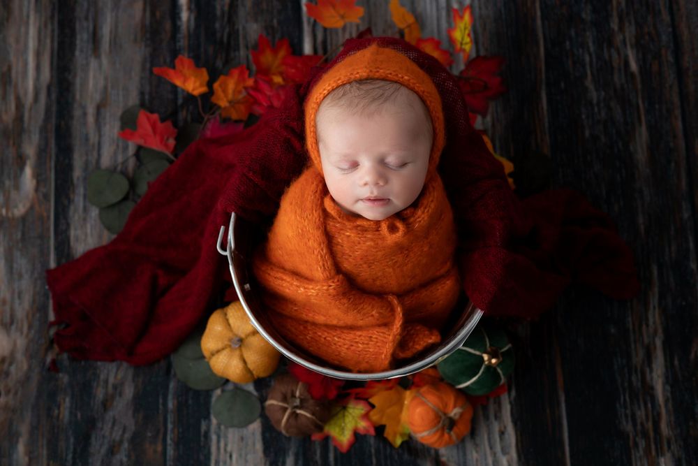 A sleeping baby wearing an orange knit pumpkin hat lies among autumn leaves and decorative fall set up.
