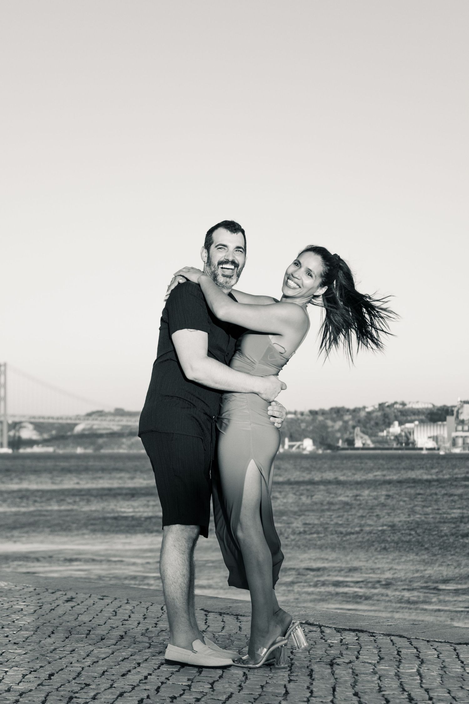 A joyful couple embraces and laughs together on a cobblestone path near the water on a sunny day.