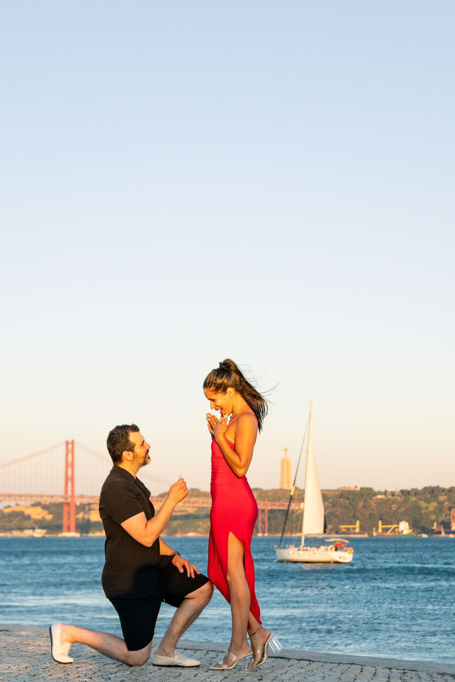 A romantic proposal scene with the Golden Gate Bridge and sailboat in the background during sunset.