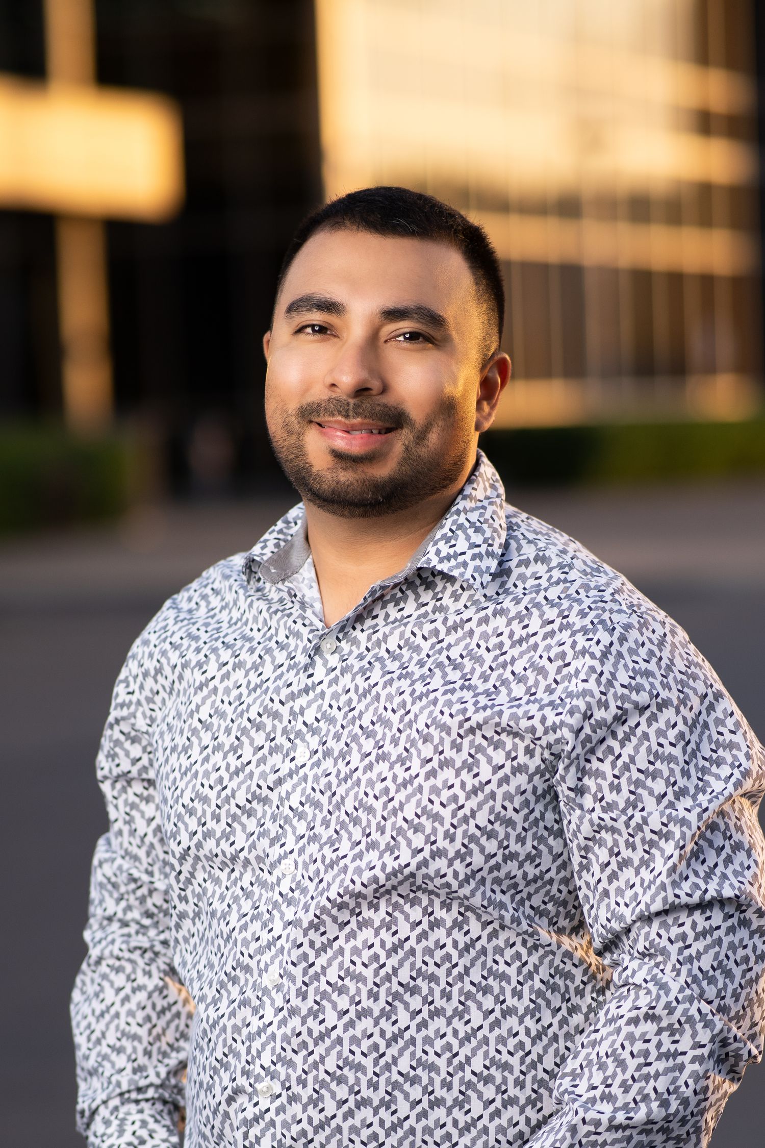 Professional headshot of a person in a dark suit jacket and blue checkered button-down shirt against a gray background.
