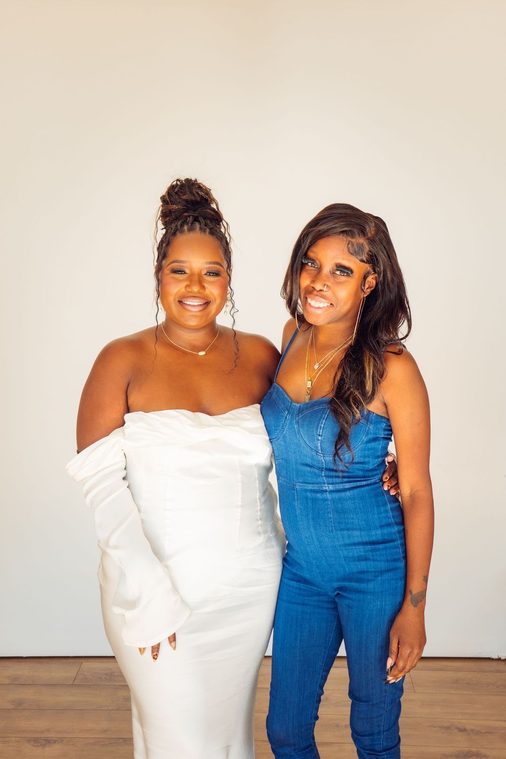 Two friends embrace while wearing a white dress and blue denim outfit for photos.