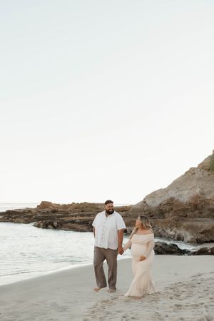 A couple walks hand in hand along a rocky beach at sunset wearing light colored clothing and creating romantic silhouettes.