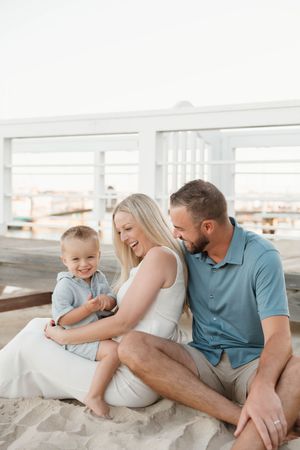Happy family of three sitting on sandy beach, parents smiling at toddler son in a bright, airy outdoor setting.