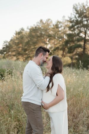 A couple shares an intimate moment in a sun-lit meadow with tall grass and pine trees in the background.