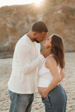 Romantic couple in white tops share an intimate moment at sunset on a beach with rocky cliffs in the background.
