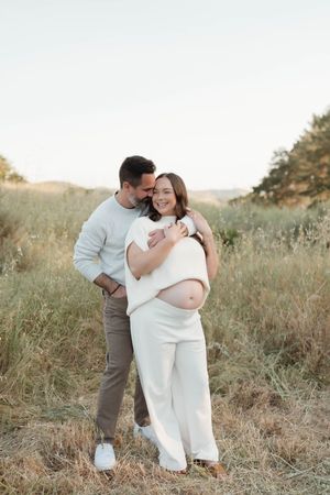 A couple embraces in a sunlit meadow during a maternity photoshoot, wearing white and neutral tones.