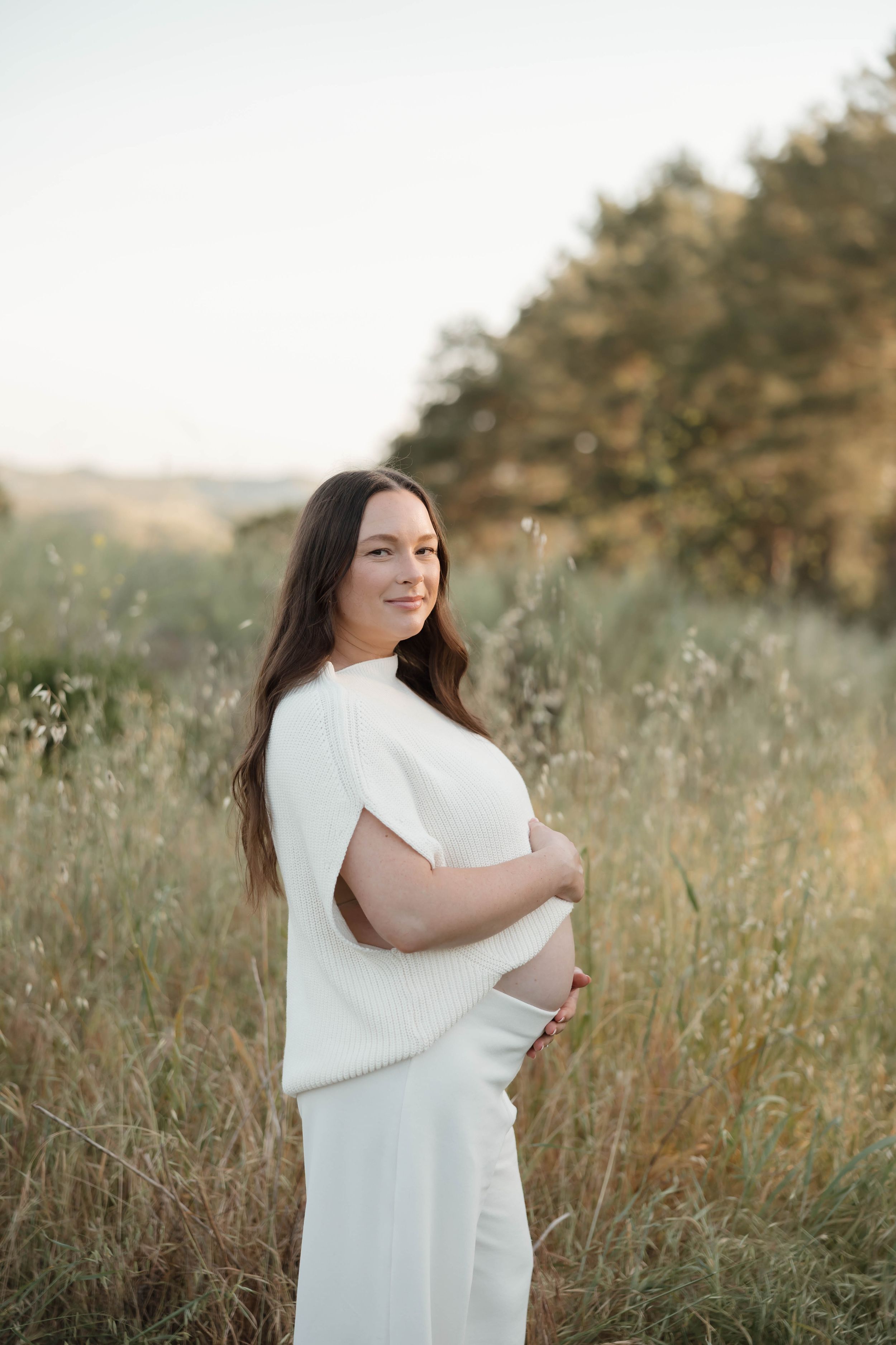 A person in a white dress stands in a sunlit meadow with tall grass and trees in the background.