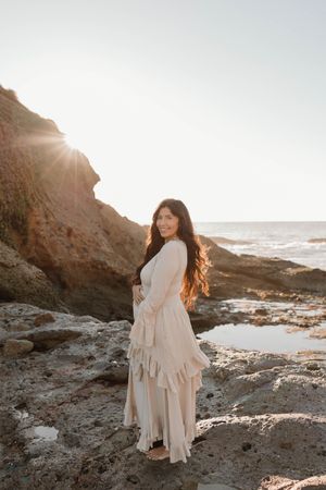 A person in a flowing cream dress stands on rocky coastal cliffs at sunset near the ocean.