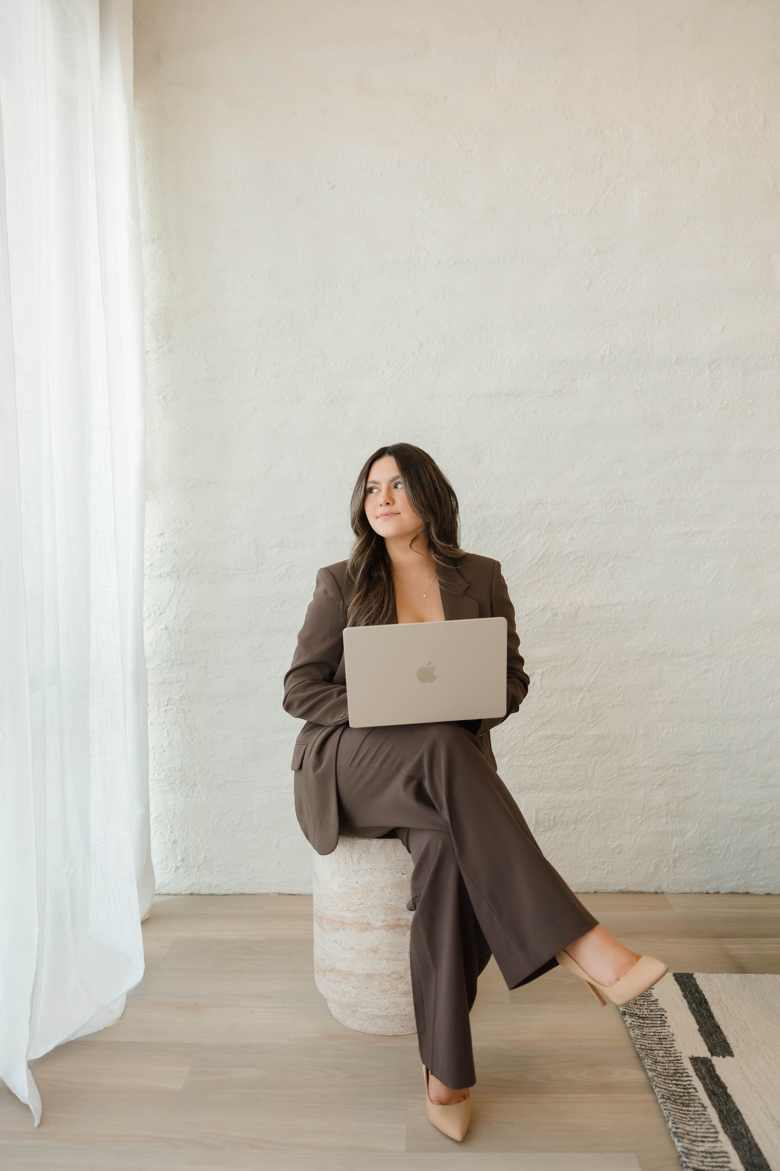 Professional sitting with laptop in minimalist white space wearing brown blazer in artistic photo series.