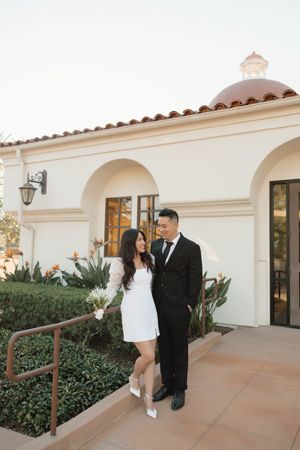 A couple in formal attire shares an intimate moment on a walkway outside a Mediterranean-style building.
